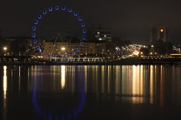 Obraz premium Night landscape with a view of the historical buildings of the city center, the Ferris wheel, evening lights and illuminations, as well as their perfect reflections in the mirror surface of the water.