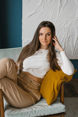 Obraz premium Beautiful young woman in beige trousers and a jumper is sitting at home in an armchair and looking at the camera