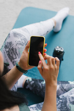 Over her shoulder, a close-up view of a sporty female yoga teacher sitting on a mat, holding a mock-up of a phone screen in her hands, watching an online workout video, using a fitness app at home