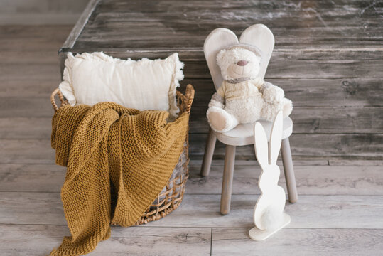 Children's soft toy bear sits on a chair, next to it there is a plywood bunny and a basket with a blanket and a pillow. Accessories in the summer room