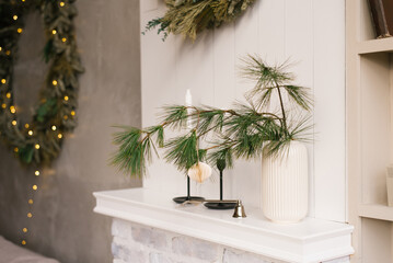 Spruce or pine branches in a white vase and candles in candlesticks on a shelf in a cozy room decorated for Christmas and New year