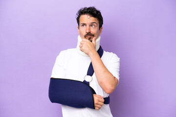 Young caucasian man wearing a sling and neck brace isolated on purple background having doubts
