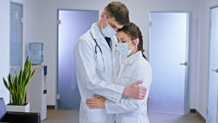 In the m of the luminous hospital corridor couple of doctors with a protective mask mask in front of the camera hugging each other they have a sad face