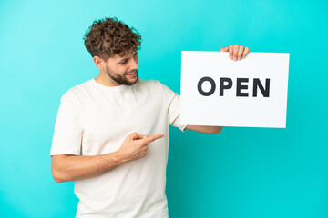 Young handsome caucasian man isolated on blue background holding a placard with text OPEN and  pointing it