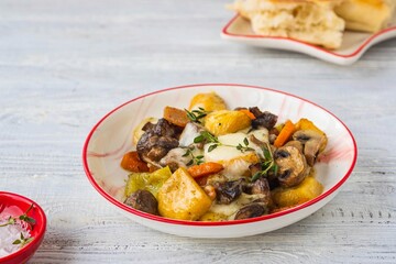 Baked vegetable mix of carrots, turnips and onions with mushrooms and cheese in a white plate on a light wooden background. Vegetable recipes.