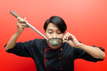 Smiling Young asian male chef in black chef uniform gesture tasting ladle standing over red isolated background. chef occupation concept. chef smelling delicious food