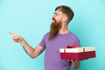 Redhead man with long beard holding a gift isolated on blue background pointing to the side to present a product