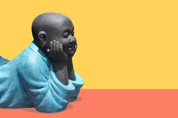 Buddha in blue clothes lies on his stomach, holding his head with his hands on a yellow-orange background. Space for text on the right.