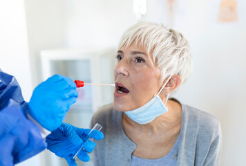 mature Caucasian woman in a clinical setting being swabbed by a healthcare worker in protective garb to determine if she has contracted the coronavirus or COVID-19.