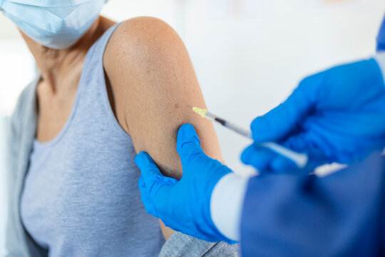 Doctor Holding Syringe Making Covid 19 Vaccination Injection Dose In Shoulder Of Female Patient Wearing Mask. Flu Influenza Vaccine Clinical Trials Concept, Corona Virus Treatment, Close Up View.