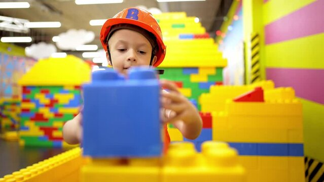 Baby boy stands in helmet and clothes of builder and makes house out of constructor bricks. Game in profession in children. Construction of toy building. Choosing future profession in childhood.