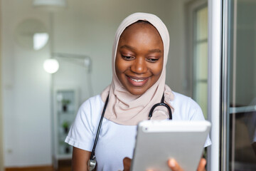 Young female musllim African clinician doctor in scrubs using touchpad while communicating with...