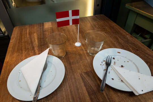 Copenhagen, Denmark  A Table In A Cafe With Two Empty Plates And Glasses And A Danish Flag.