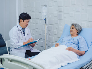 Obraz premium Smiling Asian doctor in white suit take notes while discussing happy Asian elderly, senior woman patient who lying on bed with receiving saline solution in hospital. Health care and medical concept.