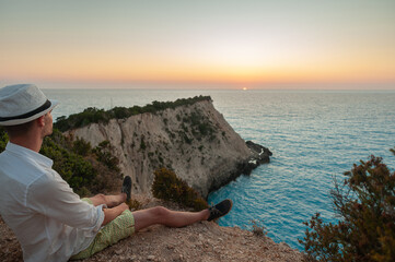 A young man is sitting on the edge of a cliff in Greece and watching the beautiful sunset