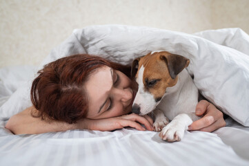 Caucasian red-haired woman sleeps in an embrace with a jack russell terrier dog on a white sheet.