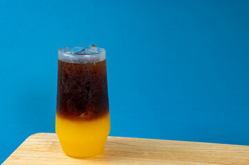 A glass of iced americano black coffee and layer of orange on a wooden table with blue background, Space for text , Orange americano.