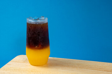 A glass of iced americano black coffee and layer of orange on a wooden table with blue background, Space for text , Orange americano.