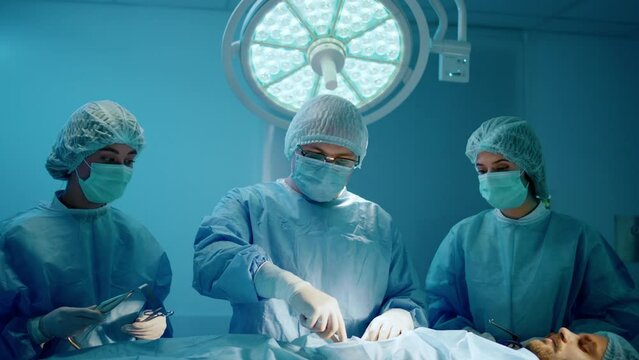A doctor and two assistant nurses are wearing scrubs and surgical gear while being in a hospital room, the doctor is performing a surgery