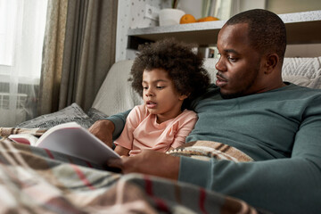 Black father and little son reading book on sofa