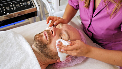 Obraz na płótnie Canvas Adult man on cosmetic procedures in the cosmetology salon. Male on face cleaning in the salon. Cosmetologist wipes a man's face with wet wipes in a spa salon.
