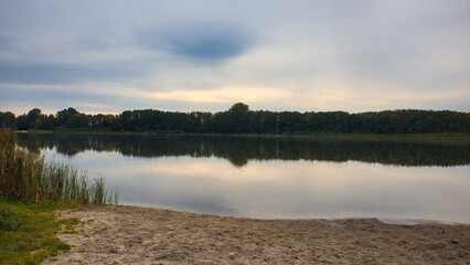 Fototapeta premium Ruhiger See mit Spiegelung des bewölkten Himmels am sandigen Ufer in der Dämmerung, Naturlandschaft am Gorinsee (Wandlitz), Landkreis Barnim, Brandenburg, Deutschland