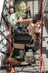 Vertical smiling anticipating student friends carrying boxes with plants, clothes and look up on stairs while relocation