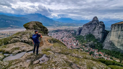 Fototapeta premium Tourist man with scenic aerial view of tourist village of Kalambaka, Thessaly, Greece, Europe, Pindus mountains. Dramatic moss overgrown rock complex of Meteora seen from Holy Trinity Monastery