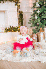 A little girl under one year old in an airy dress in a room decorated for Christmas,  near the Christmas tree among the pillows, gifts, garlands and pine needles. Christmas mood. 