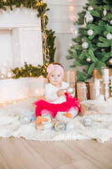 Obraz na płótnie Canvas A little girl under one year old in an airy dress in a room decorated for Christmas, near the Christmas tree among the pillows, gifts, garlands and pine needles. Christmas mood. 