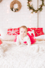 A little girl under one year old in an airy dress on a large bed in a room decorated for Christmas, among the pillows of garlands and pine needles. Christmas mood. Children and Christmas