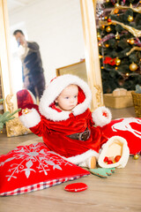 A little girl under one year old dressed as Santa Claus in a room decorated for Christmas, near the Christmas tree among the pillows, gifts, garlands and pine needles. Christmas mood. Children and Chr