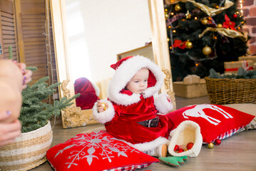 A little girl under one year old dressed as Santa Claus in a room decorated for Christmas, near the Christmas tree among the pillows, gifts, garlands and pine needles. Christmas mood. Children and Chr
