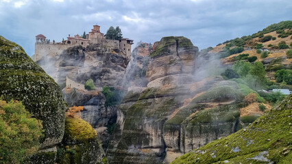 Scenic view of Holy Monastery of Varlaam on cloudy foggy day, Kalambaka, Meteora, Thessaly, Greece, Europe. Rock formations overgrown with green moss creating moody atmosphere. UNESCO World Heritage