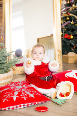 A little girl under one year old dressed as Santa Claus in a room decorated for Christmas, near the Christmas tree among the pillows, gifts, garlands and pine needles. Christmas mood. Children and Chr