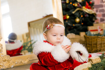 A little girl under one year old dressed as Santa Claus in a room decorated for Christmas, near the Christmas tree among the pillows, gifts, garlands and pine needles. Christmas mood. Children and Chr