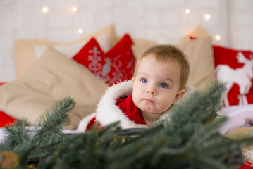 A little girl under one year old dressed as Santa Claus on a large bed in a room decorated for Christmas, among the pillows of garlands and pine needles. Christmas mood. Children and Christmas