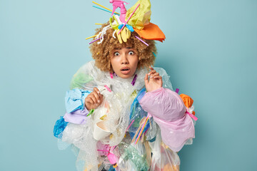 Shocked frightened woman stares at camera with worried expression made costume of plastic garbage has widely opened eyes takes care of environmental protection isolated over blue background.