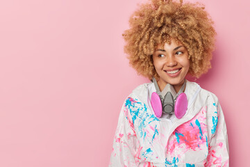 Happy curly haired woman wears biohazard chemical protective suit and gas mask looks gladfully aside smiles positively isolated over pink background with empty space for your text or advert.