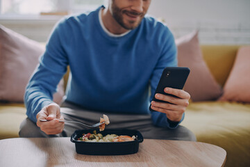 Close-up of man eating lunch and using smart phone while sitting on the couch at home