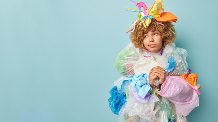 Sad thoughtful curly woman keeps hands together looks aside thinks about environmental pollution makes costume of plastic garbage poses against blue background empty space for your promotion