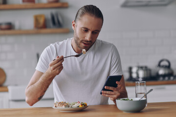 Handsome young man enjoying lunch and using smart phone while sitting at the kitchen