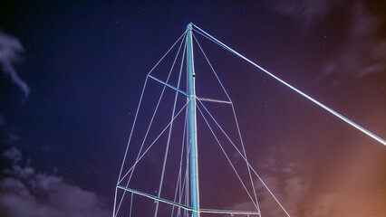 The mast of the ship against the background of the night starry sky with clouds and yellow glow