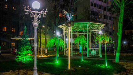 Small birch trees glow with green light from lanterns and a statue of a stork in the park