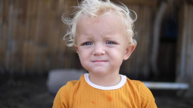 A cute blue-eyed baby stands on the street and looks at the camera. The kid is obviously dissatisfied with something, all emotions can be seen on his face.
