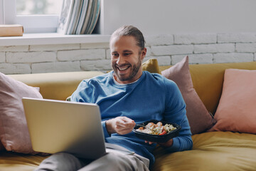 Cheerful young man eating lunch and looking at laptop while sitting on the couch at home