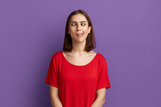 I lost my mind. Pretty brunette girl in red t-shirt demonstrating tongue and making stupid face with crossed eyes. Looks as a crazy woman showing ridiculous grimace. Posing over purple background.