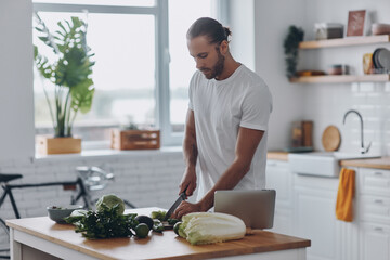 Confident young man cutting veggies while standing at the kitchen island at home