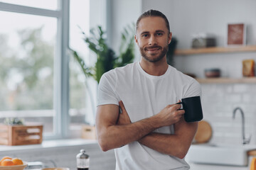 Fototapeta Handsome young man holding coffee cup and looking at camera while standing at the kitchen obraz