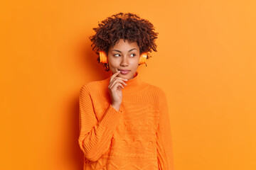 Fototapeta na wymiar Horizontal shot of beautiful thoughtful woman with curly hair keeps hand near lips looks confusingly aside dressed in jumper and headphones isolated over vivid orange background. Let me think a bit